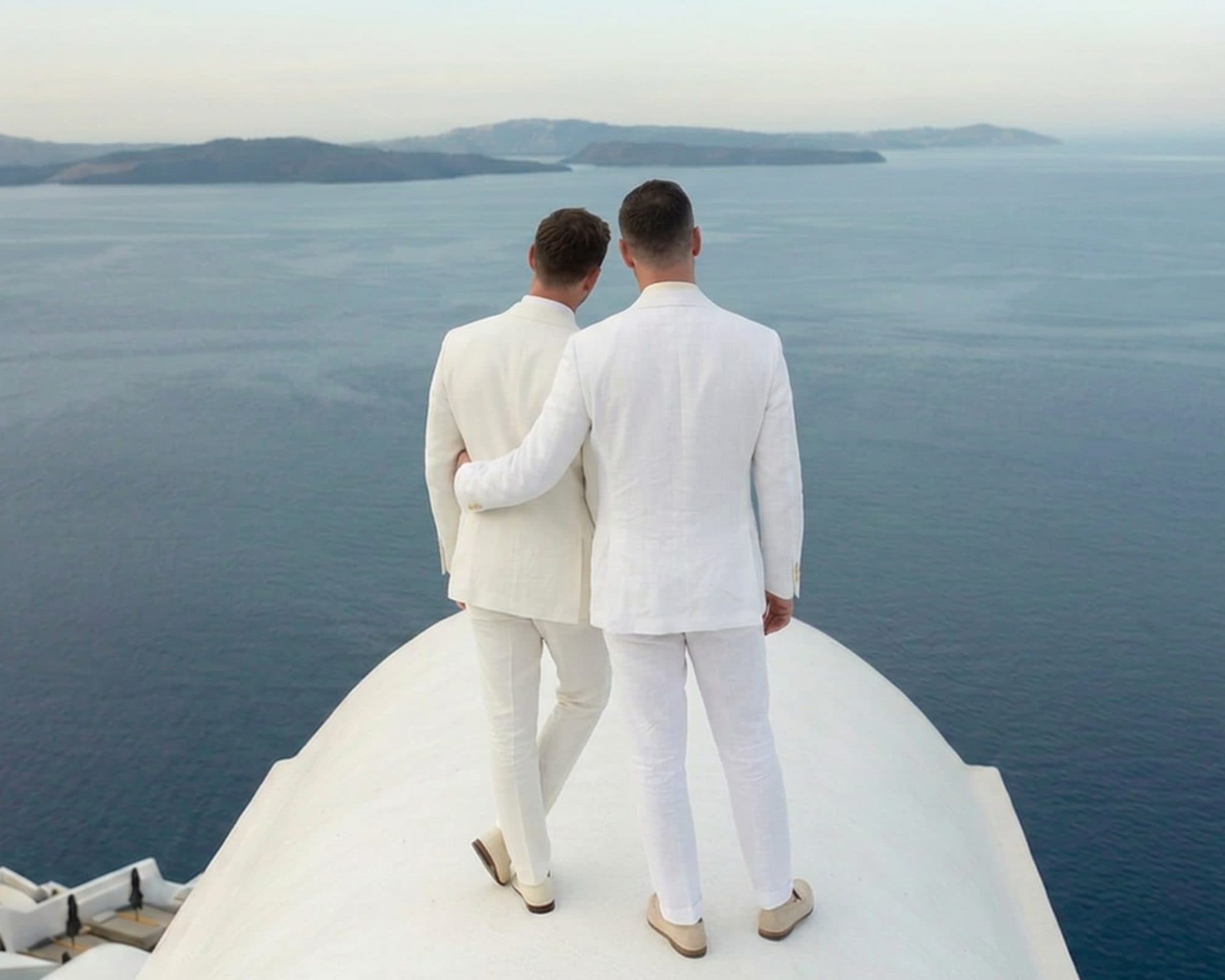Two grooms in white linen suits on a Santorini terrace above the caldera