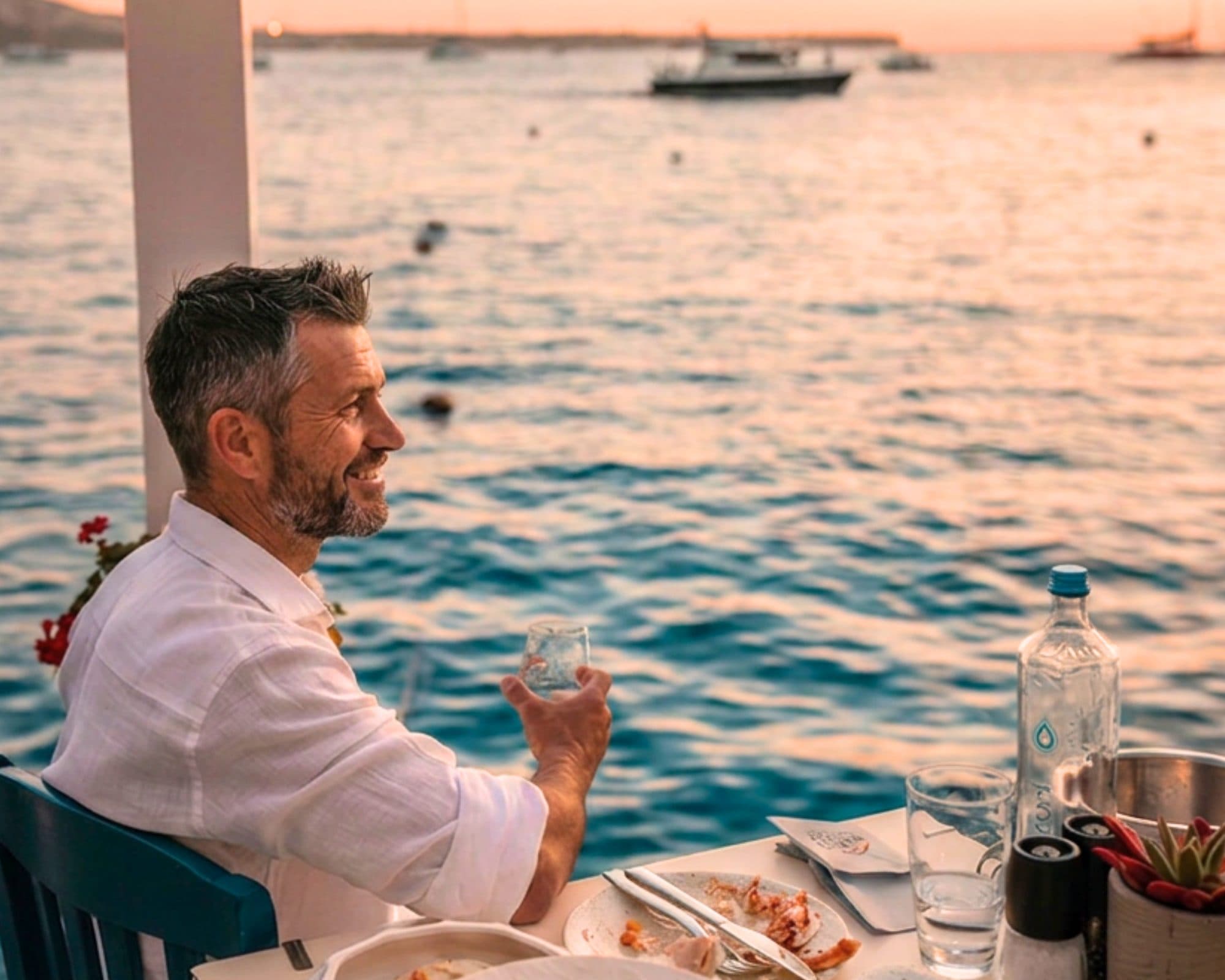 A man in his fifties at a waterside taverna table at sunset in Greece